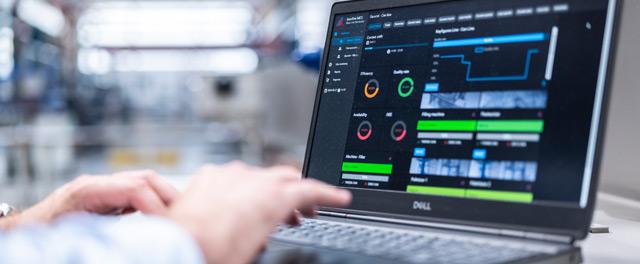 Ein Laptop zeigt Produktionsdaten und Diagramme in einer Fabrikumgebung an, im Hintergrund ist eine industrielle Maschine zu sehen.